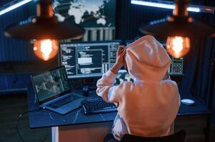 Sitting, in white hoodie. Young professional female hacker is indoors by computer with lot of information on displays photo