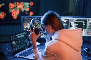 Taking ,of ,headphones. Young professional female hacker is indoors by computer with lot of information on displays photo