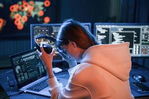 Taking of headphones. Young professional female ,hacker is indoors by computer with lot of information on displays, photo