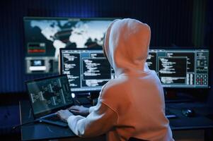 In white hoodie. Young professional female hacker is indoors by computer with lot of information on displays, photo