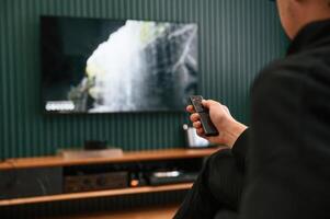 Man is sitting at home, watching TV, holding remote controller photo
