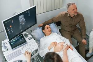 Man is sitting with ,his wife. Pregnant woman is lying down in the hospital, doctor does ultrasound photo