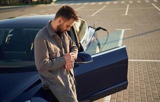 Charging the automobile. Man ,is standing, near his electric car outdoors photo