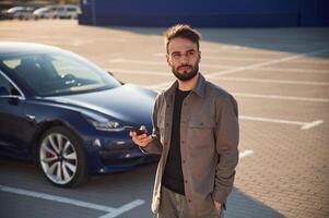 Holding smartphone in ,hand., Man is standing near his electric car outdoors photo