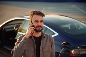 Having a call while automobile is ,charging,. Man is standing near his electric car outdoors photo