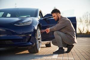 Sitting and checking the wheel. Man is standing near his electric car outdoors photo