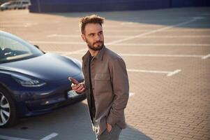 Holding smartphone ,in, hand. Man is standing near his electric car outdoors photo
