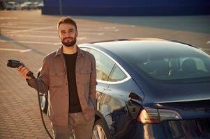 Time, to, charge the automobile. Man is standing near his electric car outdoors photo
