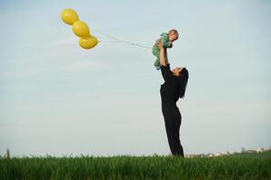 Yellow colored, balloons in hands. Mother with her little baby son is outdoors on the agricultural field photo