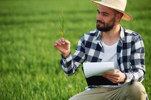 In straw hat. Holding, wheat and notepad. Handsome young man is on agricultural field photo