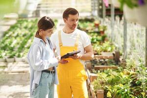 Standing, using digital tablet. Florist man and woman are working together in bright greenhouse photo