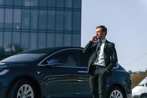 Conversation by phone. Business building behind. Man in suit and tie is standing near his car outdoors photo