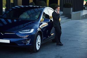 Standing, in black suit.. Businessman, is near his car outdoors photo