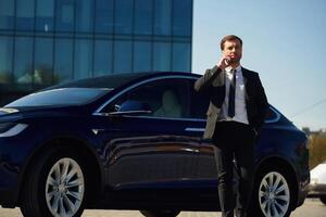 Conversation, by phone. Business building behind. Man in suit and tie is standing near his car outdoors photo