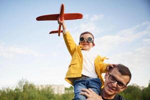 Boy is sitting on man's shoulders.. Father with his little son playing with toy plane on the field photo