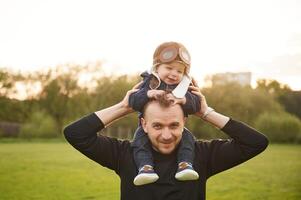Boy, is sitting ,on man's shoulders. Father with his little son playing with toy plane on the field photo