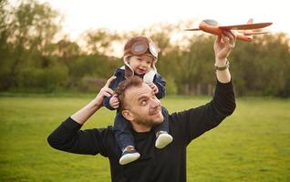 Boy is sitting on man's shoulders. ..Father with his little son playing with toy plane on the field photo