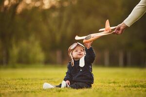 Little, boy in aviator glasses is sitting on the field. Woman's hand giving toy plane to him photo