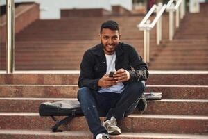 Handsome Indian student is outdoors sitting on the stairs with smartphone photo