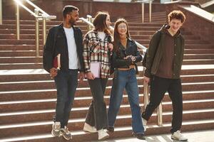 Walking, holding notepads. Four young ,,students in casual clothes are together outdoors photo