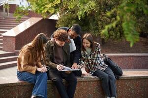 Man is holding notepad, everyone is laughing. Four young students in casual clothes are together outdoors photo