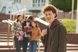 Guy with, curly hair is standing in front of his friends. Four young students in casual clothes are together outdoors photo