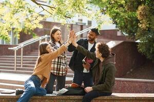 Doing, high, five. Four young students in casual clothes are together outdoors photo