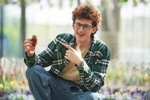 Sitting and holding the little cactus. Young man with curly hair and in glasses is in greenhouse photo