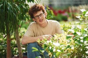 Growing ,flowers. Conception, of watering. Young man with curly hair and in glasses is in greenhouse photo