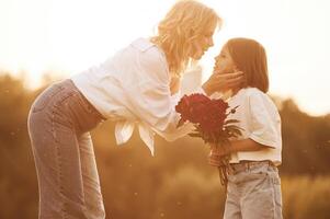 Kid is ,holding red flowers. Woman with her little daughter are on the summer field together photo