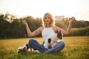 Sitting ,in yoga lotus pose. Young woman is with two little dogs on the summer field photo