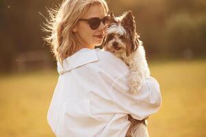 Woman ,in sunglasses is standing on the summer field and holding little dog photo