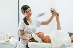 Taking a look in the mirror. Woman face getting facial care by beautician hands at spa salon photo