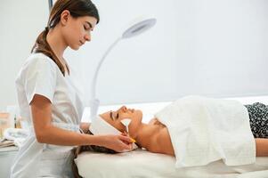 Lying down. Using ,brushes. Woman face getting facial care by beautician hands at spa salon photo