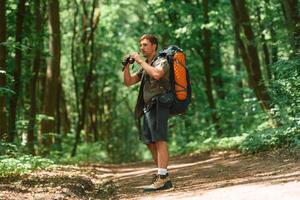 Standing and ,holding binoculars.. Tourist in summer forest. Conception of exploration and leisure photo