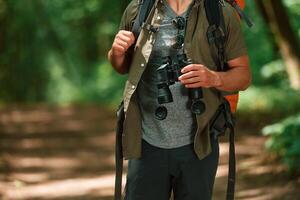 Holds, binoculars.. Close up view of tourist that is in summer forest photo