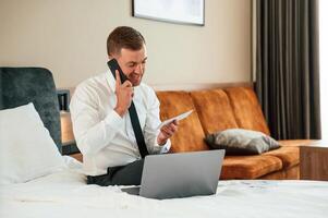 Holding tickets, sitting, on the bed ,with laptop and talking by phone. Businessman is indoors in the hotel room photo