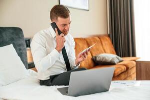 Holding tickets, sitting on the bed ,with, laptop and talking by phone. Businessman is indoors in the hotel room photo