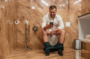 Watching funny content. A man in a white shirt is sitting on the toilet, holding a smartphone in his hands photo