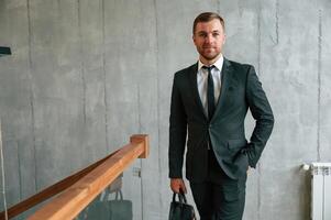 Beautiful, man in suit and tie is standing indoors and holding case in hand photo