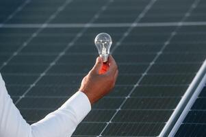 Conception of green energy. Close up view of man's hand that is holding light bult in front of solar panel photo