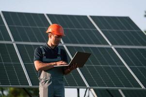 Talking by phone, holding laptop. Man is doing operating and maintenance in solar power plant. photo