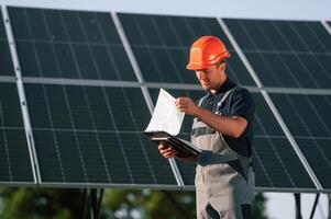 Notepad and digital tablet in hands. Man is doing operating and maintenance in solar power plant photo