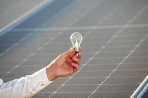 Conception of, green energy. Close up view of man's hand that is holding light bult in front of solar panel photo