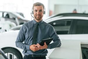 Manager is in the headphones ,and with notepad. Handsome car dealership worker is with electric vehicle photo