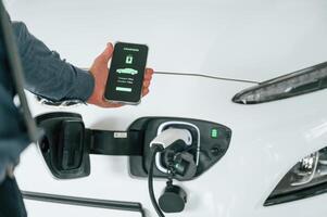 Using ,smartphone, app for monitoring charging process. Close up view of man that is with electrical car photo