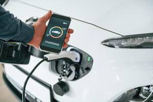 Using smartphone app for monitoring charging process. Close up view of man that is with electrical car photo