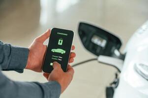 Charging process on the smartphone, app.. Close up view of man that is with electrical car photo