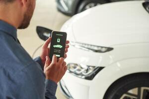 App interface of charging in smartphone. Handsome car dealership worker is with electric vehicle photo