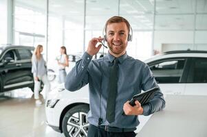 Manager, is in the headphones, and with notepad. Handsome car dealership worker is with electric vehicle photo